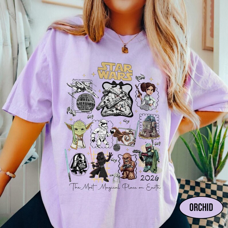 May include: A light purple t-shirt featuring a Star Wars graphic with various characters and elements from the franchise. The design includes the text "STAR WARS" and "2026 The Most Magical Place on Earth". The shirt has a relaxed fit.