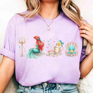 K&ouml;nnte beinhalten: Helllila T-Shirt mit einer farbenfrohen Illustration einer Meerjungfrau, einer Teekanne, einer Muschel und einem Dreizack. Die Meerjungfrau hat rotes Haar und einen t&uuml;rkisfarbenen Schwanz. Teekanne und Muschel sind mit Herzen und Sternen verziert.