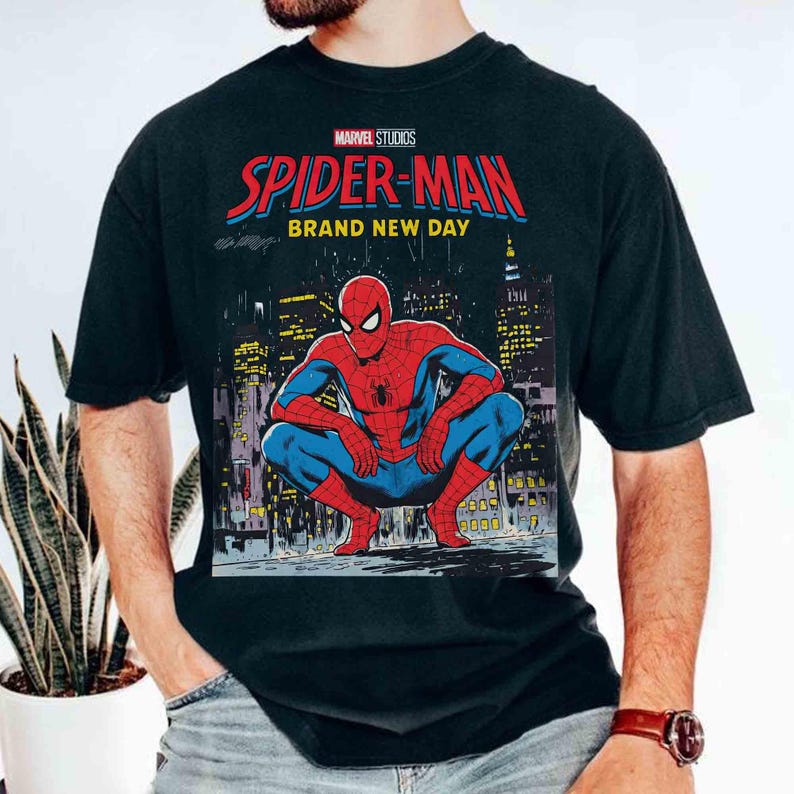 K&ouml;nnte beinhalten: Schwarzes T-Shirt mit einer Comic-Illustration von Spider-Man, der sich vor einer Stadtkulisse duckt. Der Text auf dem Shirt lautet "Marvel Studios Spider-Man Brand New Day" in Rot und Blau.