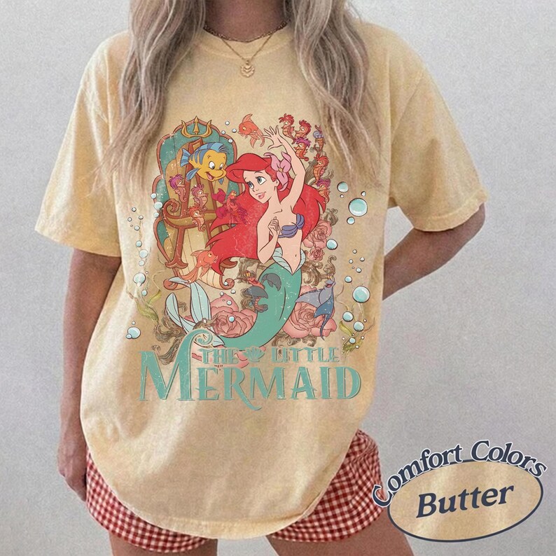 May include: A pale yellow short-sleeve t-shirt featuring a graphic print of Ariel from The Little Mermaid, surrounded by bubbles, flowers, and sea creatures. The text "THE LITTLE MERMAID" is printed in a teal serif font below the character illustration.