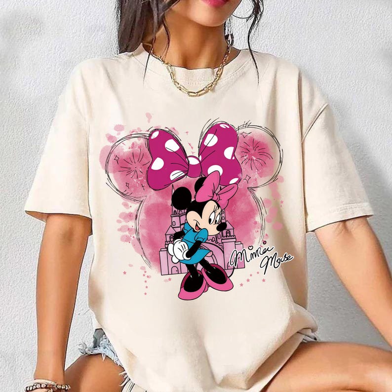 Op de afbeelding: Cr&egrave;mekleurig T-shirt met een Minnie Mouse-afbeelding. Het ontwerp bevat een roze strik met witte stippen, een roze kasteel en de tekst "Minnie Mouse" in cursief. Het shirt heeft korte mouwen.