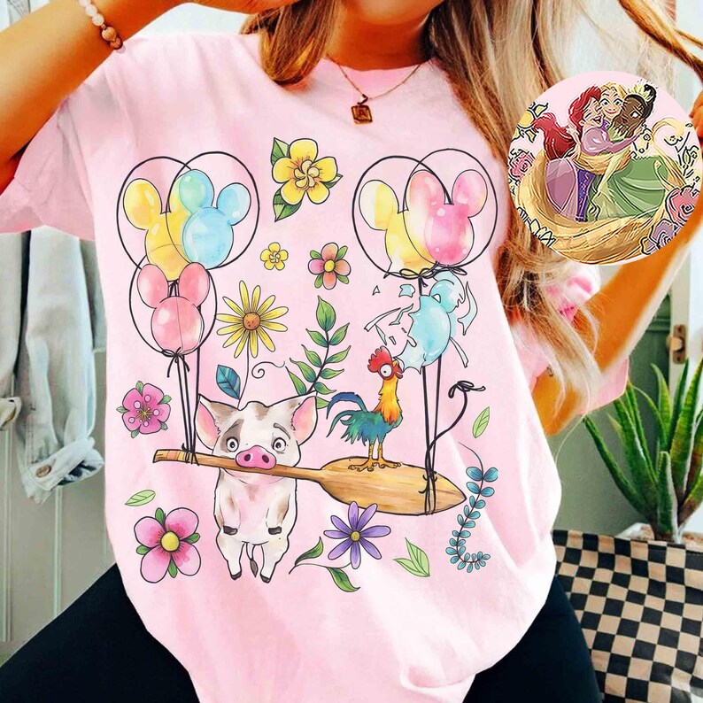 K&ouml;nnte beinhalten: Hellrosa T-Shirt mit einem farbenfrohen Aufdruck von Zeichentrickfiguren, Blumen und Luftballons. Das Design umfasst ein Schwein, das ein Holzpaddel h&auml;lt, einen Hahn und Mickey Mouse-f&ouml;rmige Luftballons. Eine runde Anstecknadel mit Zeichentrickfiguren ist ebenfalls sichtbar.