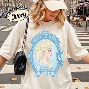 May include: Ivory-colored t-shirt featuring a light blue framed portrait of Elsa with a shield. The portrait is surrounded by a light blue decorative border. The word "ELSA" is printed on a light blue banner. The words "COMFORT COLORS" and "Ivory" are printed on the top left.