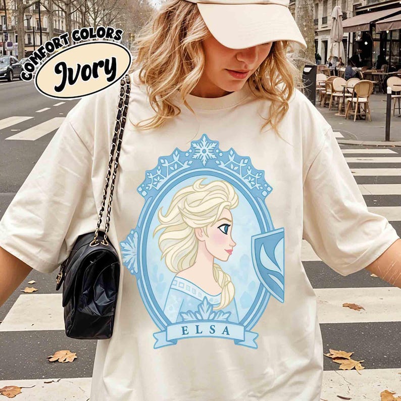 May include: Ivory-colored t-shirt featuring a light blue framed portrait of Elsa with a shield. The portrait is surrounded by a light blue decorative border. The word "ELSA" is printed on a light blue banner. The words "COMFORT COLORS" and "Ivory" are printed on the top left.