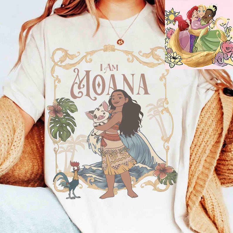 May include: Cream-colored t-shirt featuring a graphic of Moana holding a pig, with the text "I AM MOANA" in a decorative frame. The design includes tropical elements like flowers, palm trees, and a rooster. A small image of Disney princesses is in the upper right corner.