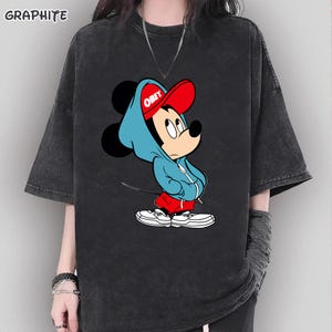 May include: Graphite-colored t-shirt featuring a cartoon Mickey Mouse character wearing a blue hoodie and red cap with the word "OBEY". The graphic tee has short sleeves and a relaxed fit.