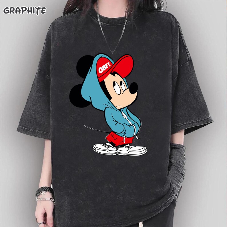 May include: Graphite-colored t-shirt featuring a cartoon Mickey Mouse character wearing a blue hoodie and red cap with the word "OBEY". The graphic tee has short sleeves and a relaxed fit.