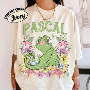 May include: Ivory-colored t-shirt featuring the name "PASCAL" in green, above a cartoon chameleon surrounded by a floral wreath. The shirt has the words "Comfort Colors" and "Ivory" in a circle.