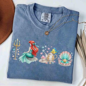 K&ouml;nnte beinhalten: Blaues Comfort Colors T-Shirt mit einem bunten Aufdruck einer Meerjungfrau, einem Dreizack und einer Muschel. Das Shirt hat einen Rundhalsausschnitt und kurze &Auml;rmel. Eine goldene Halskette mit Anh&auml;nger liegt auf dem Shirt.
