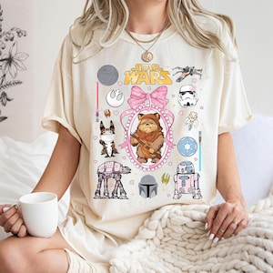 May include: Cream-colored t-shirt featuring a Star Wars-themed design. The graphic includes various characters and symbols from the franchise, such as Ewoks, R2-D2, and the Star Wars logo. The design is surrounded by a pink bow and hearts.