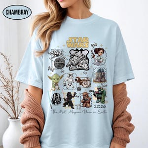 May include: Light blue t-shirt featuring a Star Wars design with various characters and elements from the franchise. The shirt includes the text "STAR WARS" and "2026 The Most Magical Place on Earth".