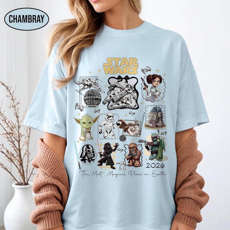 May include: Light blue t-shirt featuring a Star Wars design with various characters and elements from the franchise. The shirt includes the text "STAR WARS" and "2026 The Most Magical Place on Earth".