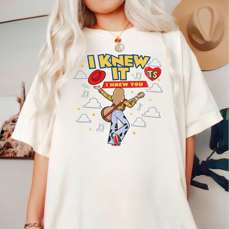 Puede incluir: Una camiseta color hueso con un estampado gr&aacute;fico de una persona de pelo largo tocando la guitarra, vistiendo una camisa de cuadros amarilla y pantalones acampanados con estampado de vaca. El texto dice I KNEW IT I KNEW YOU, con nubes y estrellas.