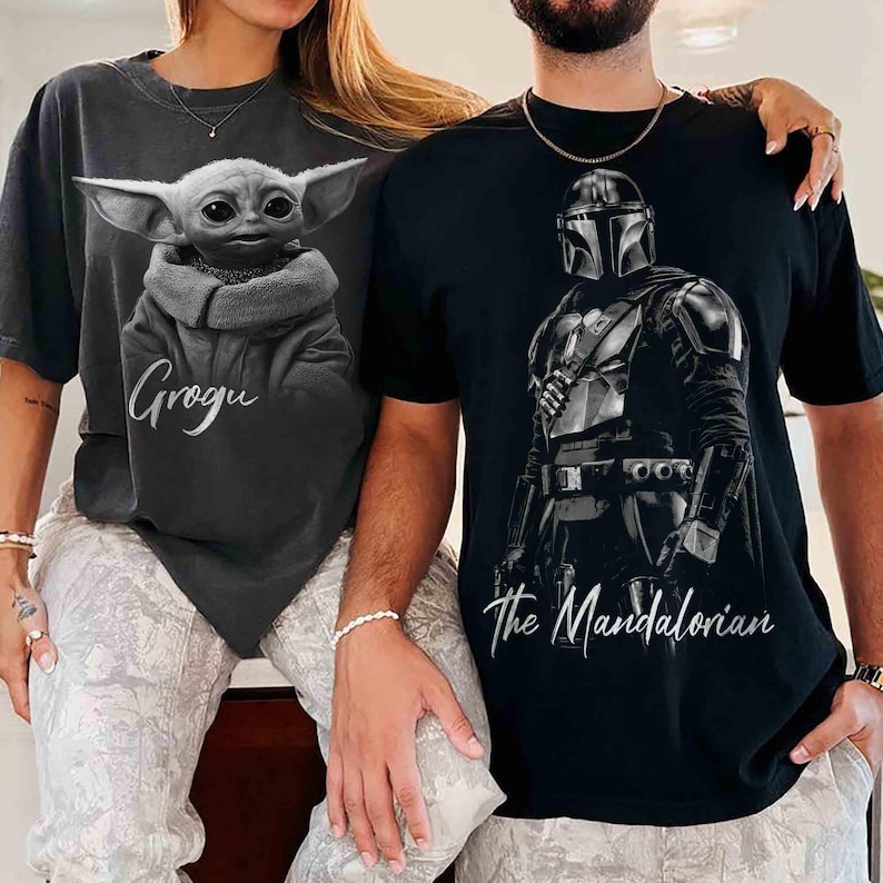May include: Two t-shirts featuring Star Wars characters. One is dark gray with Grogu's image and the name "Grogu" in script. The other is black with an image of The Mandalorian and the text "The Mandalorian".