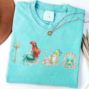 K&ouml;nnte beinhalten: Ein t&uuml;rkisfarbenes T-Shirt mit einer farbenfrohen Illustration einer Meerjungfrau, eines Dreizacks und anderer Meeresmotive. Eine goldene Halskette mit Anh&auml;nger liegt &uuml;ber dem Shirt. Das Design umfasst eine Muschel, eine Teekanne und einen Fisch.