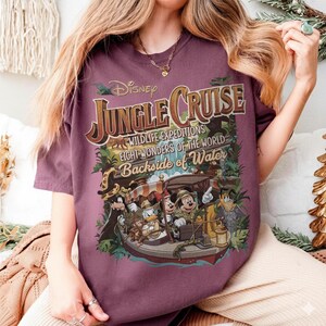 May include: A maroon t-shirt featuring a colorful graphic for Disney's Jungle Cruise. The design includes the text "Jungle Cruise," "Wildlife Expeditions," and "Eight Wonders of the World." The graphic depicts a boat with Disney characters.