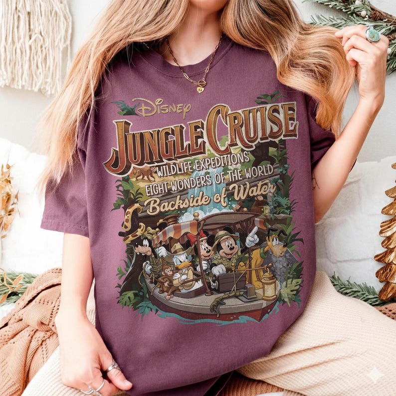 May include: A maroon t-shirt featuring a colorful graphic for Disney's Jungle Cruise. The design includes the text "Jungle Cruise," "Wildlife Expeditions," and "Eight Wonders of the World." The graphic depicts a boat with Disney characters.