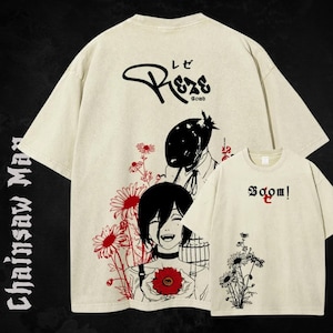 May include: Cream-colored t-shirt featuring a manga-style illustration with red and black accents. The design includes a smiling character holding a red flower, with the text "Reze Bomb" on the back and "Boom!" on a smaller shirt. The shirt is part of the "Chainsaw Man" collection.