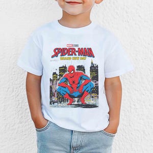 K&ouml;nnte beinhalten: Wei&szlig;es T-Shirt mit einer Grafik von Spider-Man, der vor einer Stadtkulisse hockt. Das Design enth&auml;lt den Text "MARVEL STUDIOS SPIDER-MAN BRAND NEW DAY" in Rot und Blau.