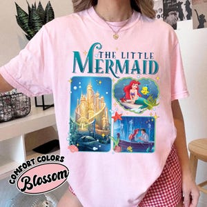 May include: A light pink t-shirt featuring the text "THE LITTLE MERMAID" in teal and white. The shirt has three graphic images: a castle, Ariel and Flounder, and Ariel and Eric in a boat. The text "COMFORT COLORS Blossom" is also on the shirt.