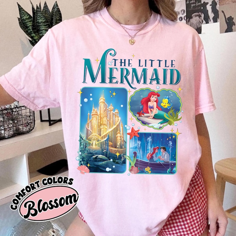 May include: A light pink t-shirt featuring the text "THE LITTLE MERMAID" in teal and white. The shirt has three graphic images: a castle, Ariel and Flounder, and Ariel and Eric in a boat. The text "COMFORT COLORS Blossom" is also on the shirt.