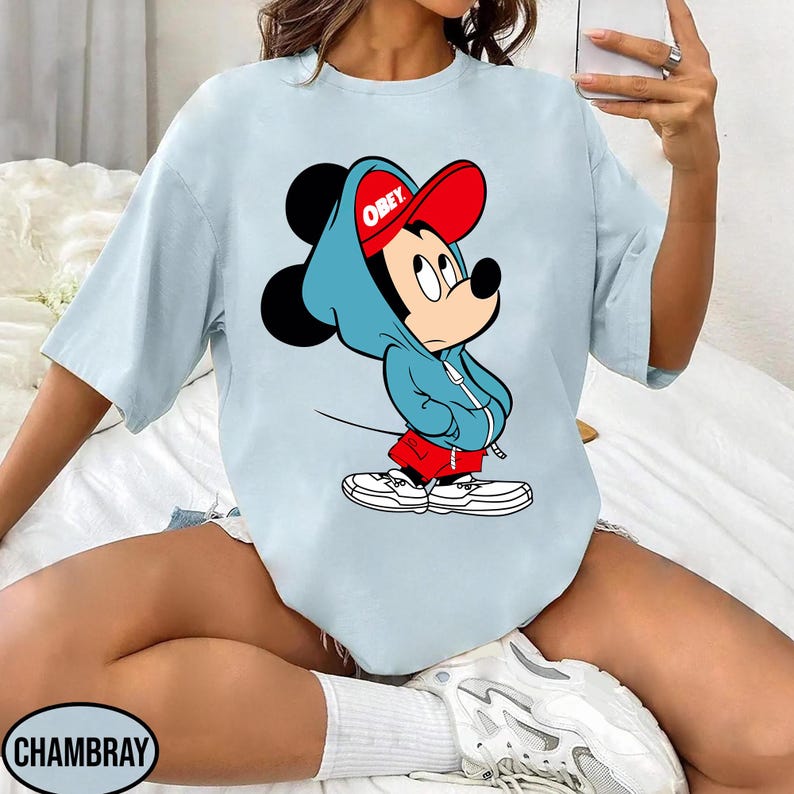 May include: Light blue t-shirt featuring a graphic of Mickey Mouse wearing a blue hoodie and a red cap with the word "OBEY". The cartoon character is also wearing red shorts and white sneakers. The shirt is a casual style.