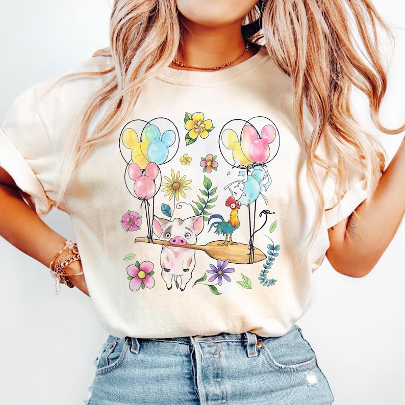 K&ouml;nnte beinhalten: Cremefarbenes T-Shirt mit einem farbenfrohen Aufdruck eines Schweins, das ein Holzpaddel mit einem Hahn, Luftballons und Blumen tr&auml;gt. Die Ballons sind wie Mickey Mouse-K&ouml;pfe geformt. Das Design umfasst verschiedene Blumen und Bl&auml;tter.