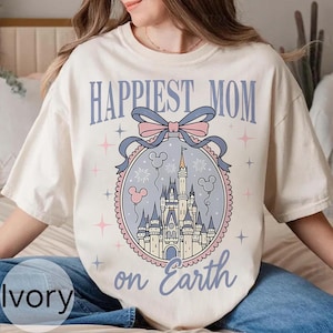 以下が含まれることがあります： アイボリー色のTシャツ。テキスト「Happiest Mom on Earth」は薄い青色の筆記体。デザインは、ピンクと青のリボン、ミッキーマウスの風船、花火のある円形のフレーム内の城のイラストが特徴です。