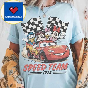 Pu&ograve; includere: T-shirt azzurra con una grafica di Topolino, Paperino, Paperina e Minnie in un'auto da corsa rossa. Il testo recita "SPEED TEAM 1928". Due bandiere a scacchi bianche e nere sullo sfondo.