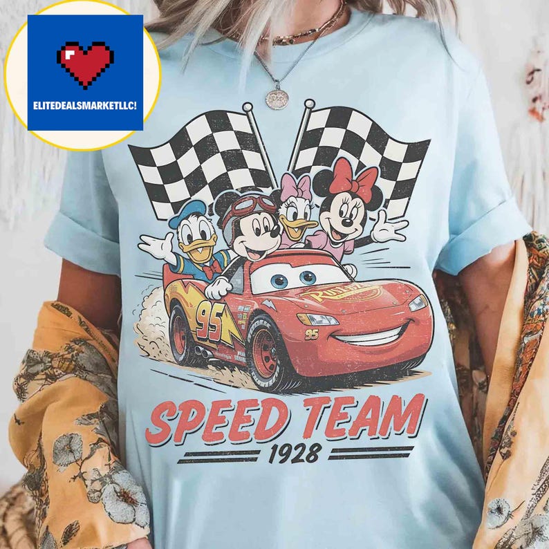 Pu&ograve; includere: T-shirt azzurra con una grafica di Topolino, Paperino, Paperina e Minnie in un'auto da corsa rossa. Il testo recita "SPEED TEAM 1928". Due bandiere a scacchi bianche e nere sullo sfondo.
