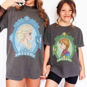 May include: Two gray t-shirts featuring Disney's Frozen characters Elsa and Anna. Each shirt has a portrait of the character in a decorative frame with their name below. The shirts are worn by two people.