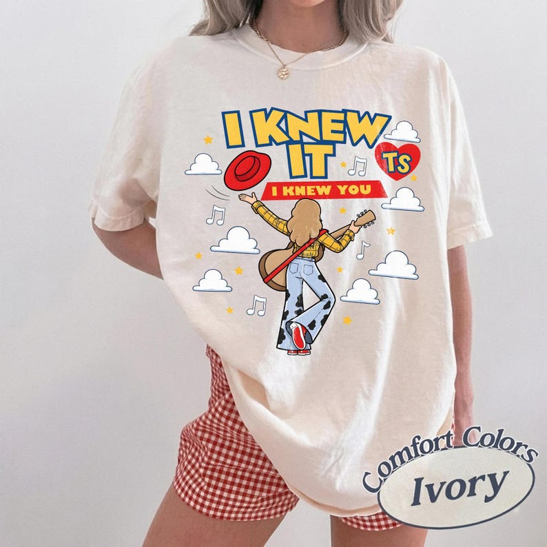 Puede incluir: Una camiseta de color marfil con un gr&aacute;fico de una persona de pelo rubio tocando la guitarra, con una camisa de cuadros amarilla y pantalones con estampado de vaca. El texto dice I KNEW IT y I KNEW YOU, junto a un sombrero rojo y nubes.