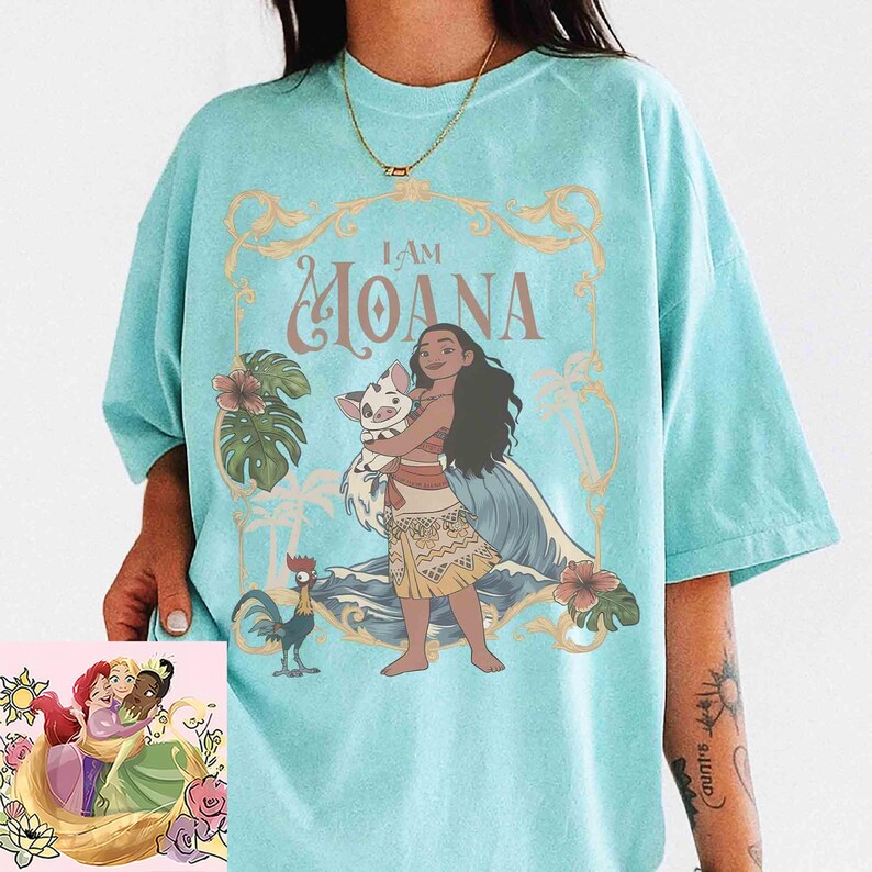 May include: A light blue t-shirt featuring the words "I AM MOANA" in a decorative font, with an illustration of Moana holding a pig. The shirt also includes a rooster and floral accents. A smaller image of Disney princesses is in the corner.
