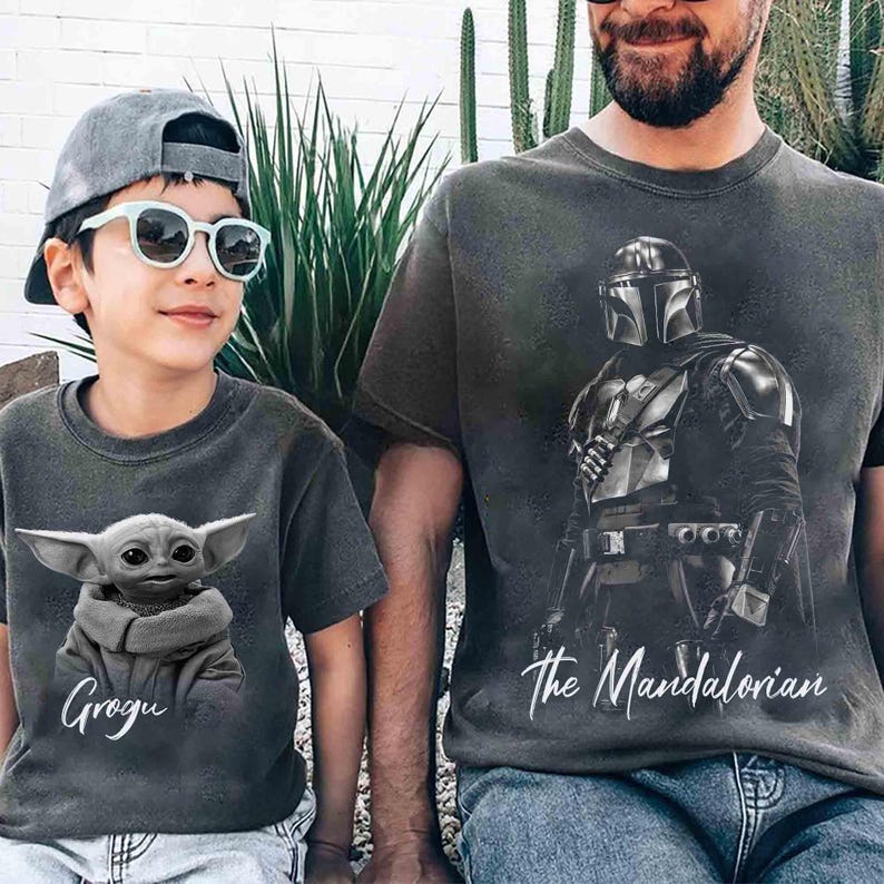 May include: Two gray t-shirts featuring characters from The Mandalorian. One shirt displays Grogu with the name "Grogu" in white script. The other shirt features The Mandalorian with the text "The Mandalorian".