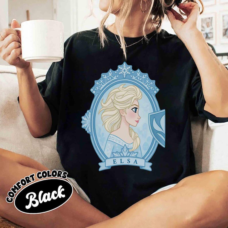 May include: Black t-shirt featuring a light blue oval portrait of Elsa from Frozen, with the name "ELSA" below. The shirt has a Comfort Colors label that says "Black".