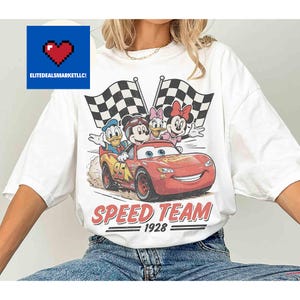 Pu&ograve; includere: T-shirt bianco con grafica di personaggi Disney su un'auto da corsa rossa. Il design include Topolino, Paperino, Paperina e Minnie, con bandiere a scacchi e la scritta "SPEED TEAM 1928".