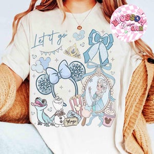 Może przedstawiać: Kremowa koszulka z jasnoniebieskim nadrukiem graficznym i napisem "Let it go". Projekt zawiera ilustracje Elsy, Olafa, uszu Myszki Miki i innych element&oacute;w tematycznych z Krainy Lodu. R&oacute;żowe i białe logo znajduje się w prawym g&oacute;rnym rogu.