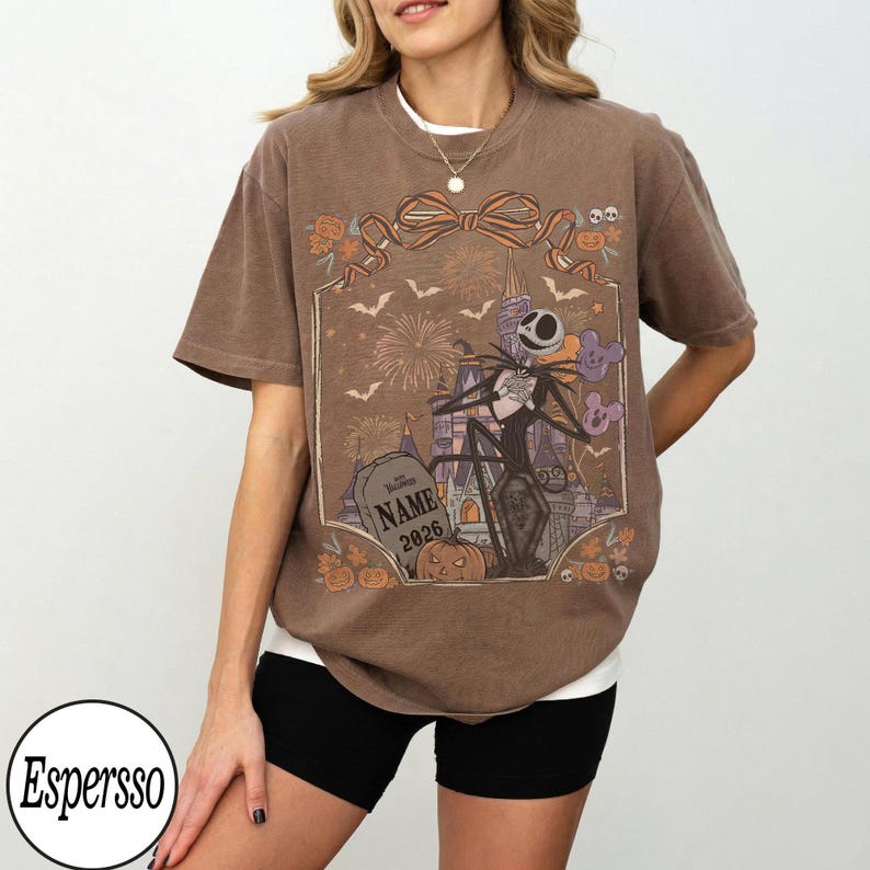 May include: A brown short-sleeve t-shirt featuring a graphic print of a skeleton character, a castle, fireworks, bats, and pumpkins. The design includes a tombstone with the text NAME 2026 and a large bow at the top of the rectangular frame.
