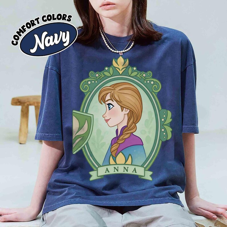 May include: Navy blue t-shirt featuring a cartoon illustration of Anna from Frozen. The design is framed in a green oval with floral accents and the name "ANNA" below. The shirt has short sleeves and the words "COMFORT COLORS Navy" are printed on the top left.