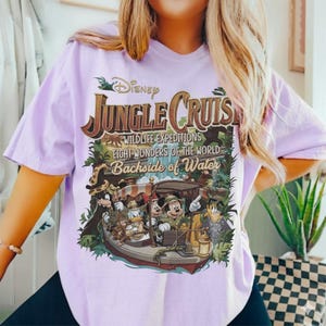 May include: A light purple t-shirt featuring a graphic for Disney's Jungle Cruise. The design includes the text "Jungle Cruise," "Wildlife Expeditions," and "Backside of Water," along with illustrations of Mickey Mouse, Donald Duck, and other characters on a boat.