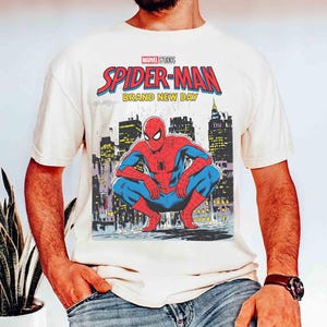 K&ouml;nnte beinhalten: Cremefarbenes T-Shirt mit einem Marvel Studios Spider-Man-Motiv. Das Design zeigt die Worte "SPIDER-MAN BRAND NEW DAY" in Rot und Blau sowie eine Illustration von Spider-Man, der sich vor einer Skyline hockt.