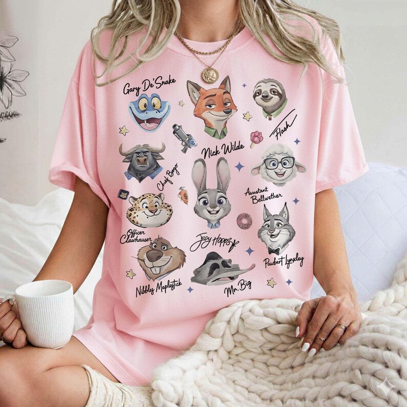 K&ouml;nnte beinhalten: Ein hellrosa T-Shirt mit Cartoon-Charakterportr&auml;ts aus dem Film Zoomania. Das Shirt zeigt Charaktere wie Nick Wilde, Judy Hopps und Flash, mit ihren Namen darunter. Das Shirt hat eine l&auml;ssige, &uuml;bergro&szlig;e Passform.