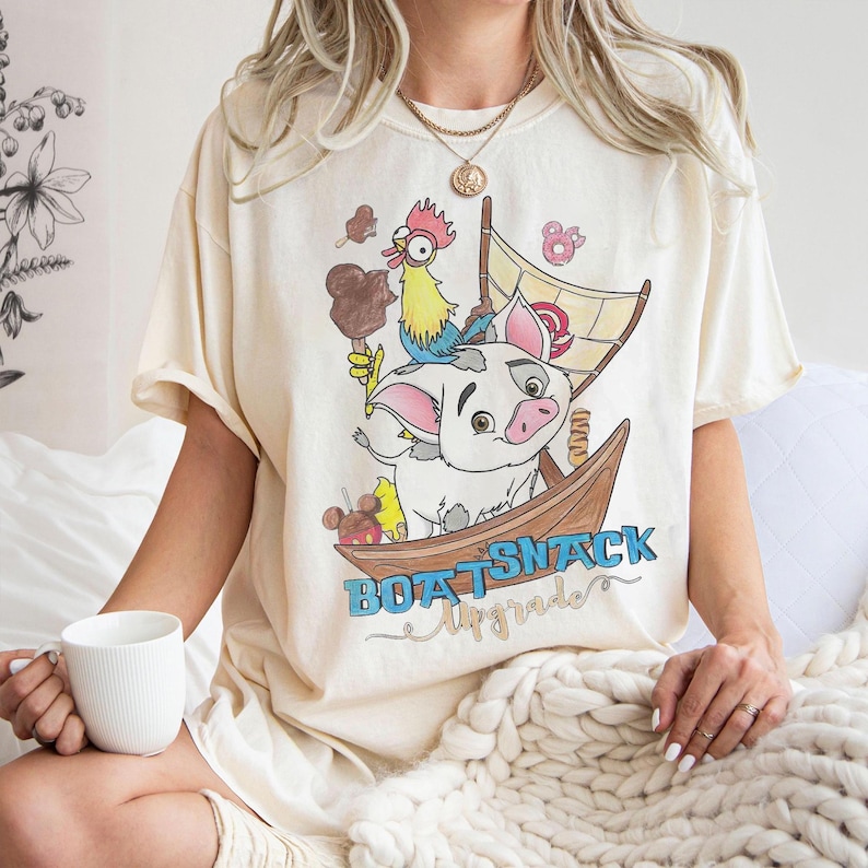 May include: Cream-colored t-shirt featuring a cartoon illustration of a pig, a rooster, and a boat with the text "BOATSNACK Upgrade". The design includes ice cream and other food items. The shirt has short sleeves.