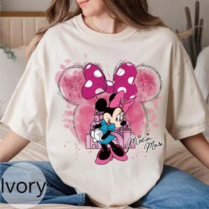 Op de afbeelding: Ivoor kleurig T-shirt met een Minnie Mouse afbeelding. Het ontwerp bevat een roze strik met witte stippen, een cartoon Minnie Mouse en een roze kasteel. De woorden "Minnie Mouse" zijn cursief geschreven.