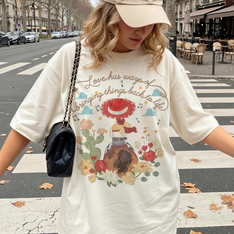 May include: An off-white t-shirt featuring a graphic of a cowgirl on a horse surrounded by cacti and flowers. The text reads, Love has ways of bringing things back to life. The design includes musical notes, clouds, and a red hat. A black quilted bag with a gold chain strap is worn over the shoulder.