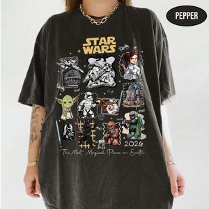 May include: Dark gray t-shirt featuring a Star Wars design. The shirt displays various characters and elements from the Star Wars franchise, including Yoda, Darth Vader, and the Millennium Falcon. The text "The Most Magical Place on Earth" is also present.