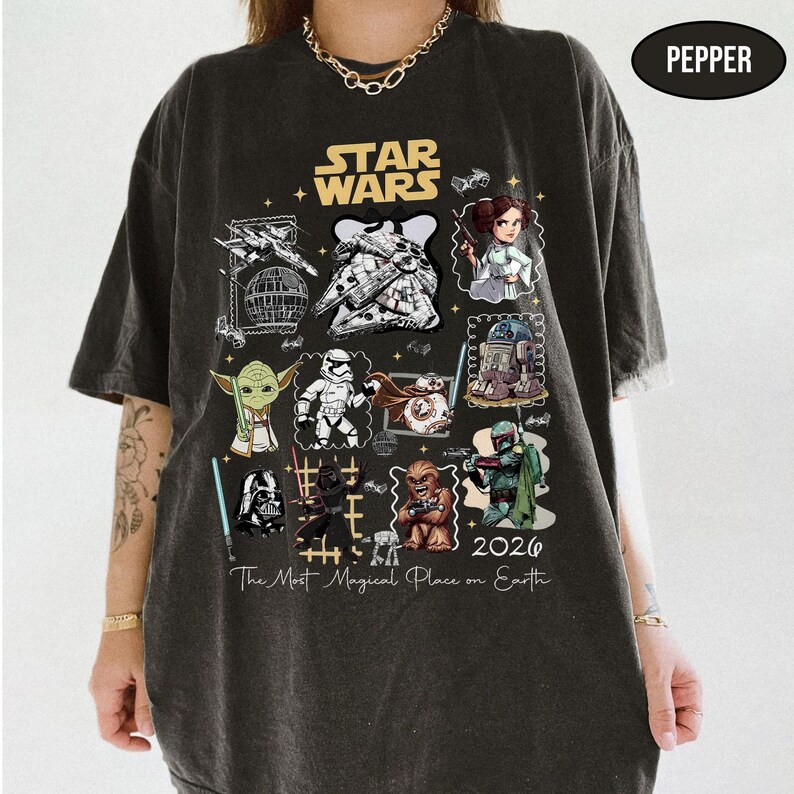 May include: Dark gray t-shirt featuring a Star Wars design. The shirt displays various characters and elements from the Star Wars franchise, including Yoda, Darth Vader, and the Millennium Falcon. The text "The Most Magical Place on Earth" is also present.