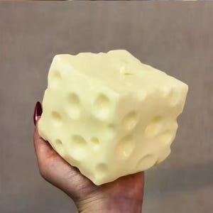 Pode incluir: Uma vela grande, amarela, em forma de cubo com numerosos buracos, assemelhando-se a um bloco de queijo suíço. A vela é segurada em uma mão com um fundo neutro. A cor da vela é amarelo pálido.
