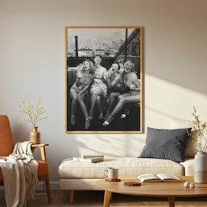 May include: A framed black and white photograph depicts five women laughing joyfully on a rooftop, with a cityscape in the background. The photograph is displayed in a room with a neutral color palette, featuring a sofa, chair, and coffee table.