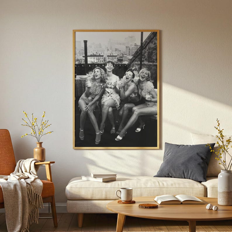 May include: A framed black and white photograph depicts five women laughing joyfully on a rooftop, with a cityscape in the background. The photograph is displayed in a room with a neutral color palette, featuring a sofa, chair, and coffee table.